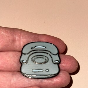 May include: A silver enamel pin depicting a retro telephone design. The pin features a silver outline and white highlights. The pin is held in a hand against a pale pink backdrop.
