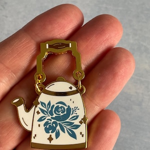 Puede incluir: Un broche de tetera de metal dorado con cuerpo blanco y diseño floral azul. El broche tiene un asa y un pico dorados. El broche se sostiene en una mano.
