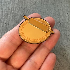 May include: A gold-toned metal enamel pin featuring a circular pastry board with a decorative leaf border and a rolling pin placed diagonally across the centre. The design has a smooth finish with dark outlines defining the shapes of the kitchen tools.