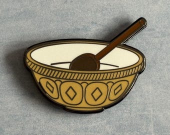 Puede incluir: Pin con un cuenco de avena con una cuchara. El cuenco es de color canela con un patrón decorativo y un contorno negro. La avena es blanca y la cuchara es marrón.