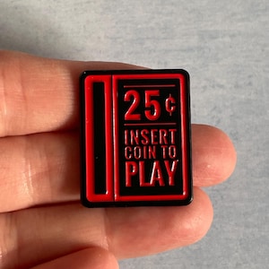 May include: A rectangular pin with a black border and a red design. The pin features the text "25¢ INSERT COIN TO PLAY" in red. The pin is a nostalgic nod to classic arcade games.
