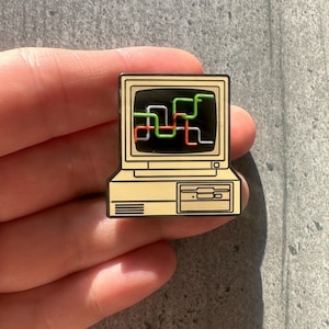 Könnte beinhalten: Ein beigefarbener Emaille-Pin in Form eines Vintage-Desktop-Computers mit einem schwarzen Bildschirm, der eine Grafik aus miteinander verbundenen grünen, orangefarbenen und weißen Linien zeigt. Das Gehäuse hat einen Laufwerksschlitz und Lautsprecher.
