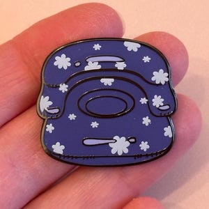 May include: A purple enamel pin shaped like a vintage telephone. The pin is decorated with white flower motifs. The pin has a black border and is held in a person's hand.