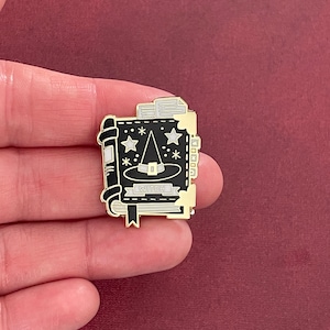 May include: Black and gold enamel pin depicting a closed book with a witch's hat and stars on the cover. The book has a gold ribbon bookmark and the text "WITCH" on the cover.