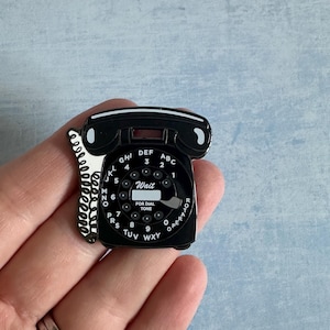 May include: A black and white rotary phone pin. The pin depicts a classic rotary dial with numbers and letters. The phone has a handset and a coiled cord. The text "Wait" and "For Dial Tone" are also visible on the pin.
