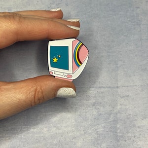 May include: A white and pink enamel pin shaped like a retro computer with a blue screen and yellow stars. The computer has a rainbow stripe on the side.