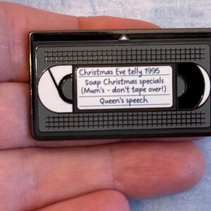 May include: A black and grey enamel pin badge shaped like a VHS cassette. The pin has a white label with the text "Christmas Eve telly 1995, Soap Christmas specials (Mum's - don't tape over!), Queen's speech."
