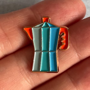 May include: A colorful enamel pin in the shape of a stovetop espresso maker. The pin features a gold outline and is filled with shades of blue, teal, orange, and gray. The handle and top are orange, while the body is a mix of blue and gray.