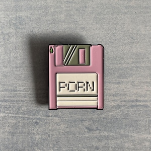 May include: A pink floppy disk-shaped pin with the word "PORN" in white block letters. The pin has a black border and a green and grey design on the top. The background is a light blue-grey.