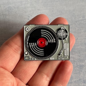 May include: A grey and black Technics SL-1200 turntable pin with a red centre. The pin features a detailed design of the turntable's components, including the tonearm and platter. The word "Technics" is printed on the bottom right.