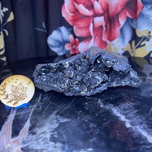 May include: A dark, textured, irregularly shaped black mineral specimen. Beside it is a small, round wooden coaster with a crystal design, a crescent moon, and a sprig of leaves. The coaster is supported by a small blue stand.
