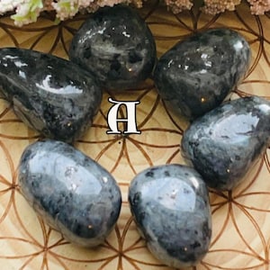 May include: Six polished labradorite stones in shades of grey and black are arranged in a circular pattern. The stones have a smooth, glossy surface. The letter "A" is visible in the centre of the arrangement. The stones are on a wooden surface.