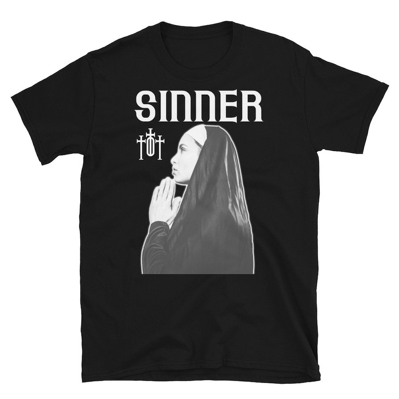 Op de afbeelding: Zwart T-shirt met het woord "SINNER" in witte tekst en een zwart-witte afbeelding van een non die bidt met twee kruisen boven haar hoofd.