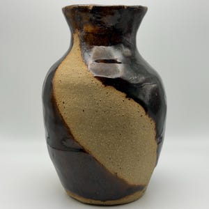 May include: A handcrafted ceramic vase with a unique design. The vase features a dark brown, glossy glaze that contrasts with a tan, unglazed section. The vase has a rounded body and a slightly flared rim, perfect for displaying flowers or as a decorative piece.