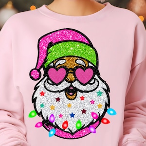 May include: Pink sweatshirt featuring a glittery Santa Claus design. Santa wears heart-shaped sunglasses, a green and pink hat, and a beard adorned with colorful stars and Christmas lights. The design has a festive, sparkly appearance.