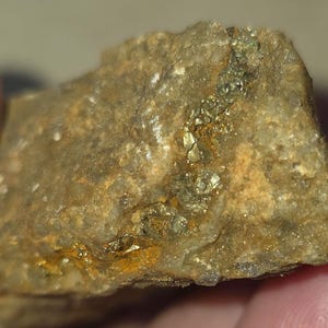 May include: A close-up of a rough, tan-coloured rock with a vein of sparkling, metallic gold-coloured minerals running through it. The rock has a textured surface and appears to be a natural mineral specimen. The overall colour palette is earthy, with shades of brown and gold.