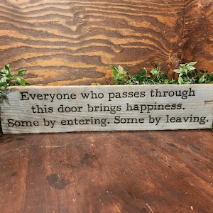 May include: A rustic wooden sign with a whitewashed background and black lettering. The sign reads "Everyone who passes through this door brings happiness. Some by entering. Some by leaving."
