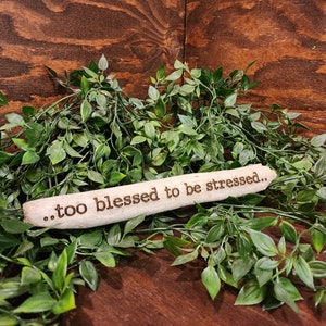 Może przedstawiać: Drewniany znak z tekstem "...too blessed to be stressed..." leżący na zielonych liściach.