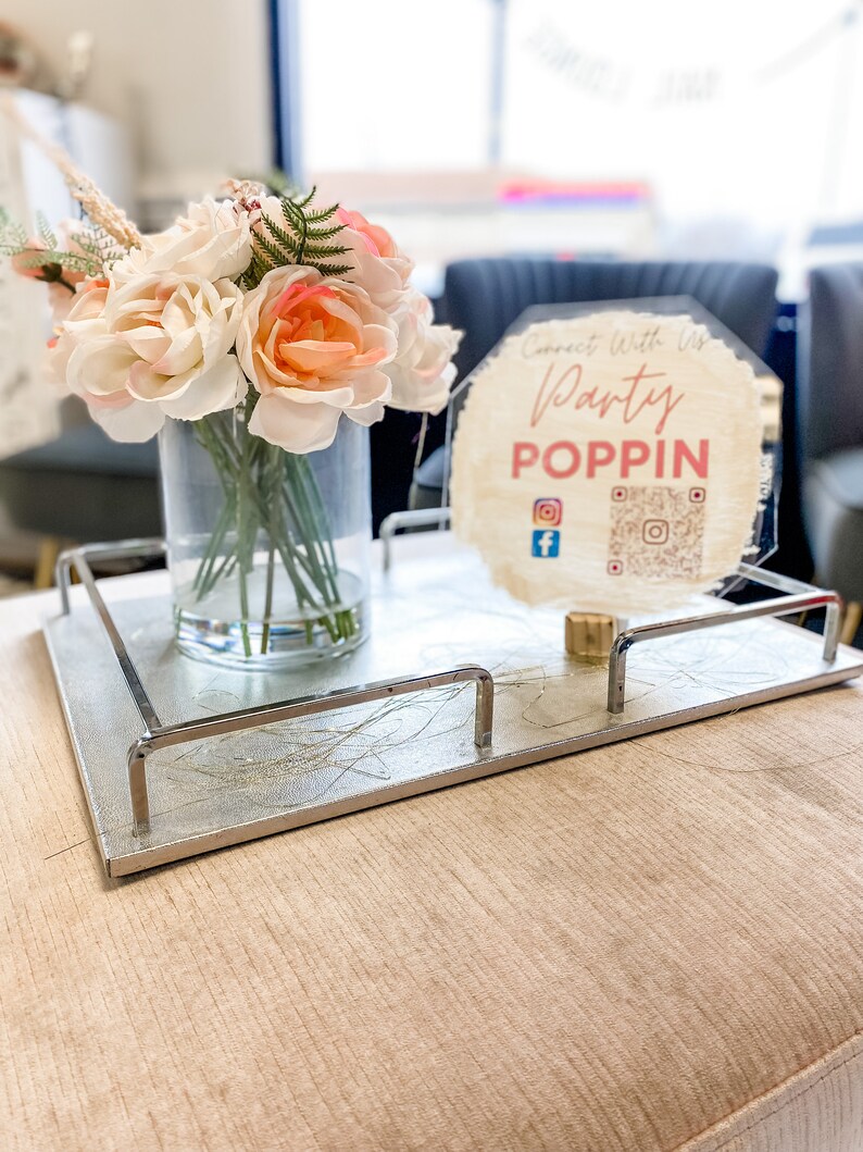 May include: A silver tray with a decorative design and metal handles, holding a clear glass vase with a bouquet of peach and white artificial flowers. A hexagonal sign with the text "Connect with us Party POPPIN" and social media icons is on the tray.