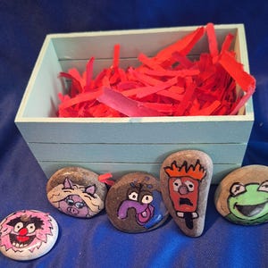 May include: A light blue wooden crate filled with red shredded paper filler, accompanied by five smooth river stones hand-painted with colorful cartoon character faces, including a pig, a purple creature, an orange-haired figure, and a green frog.