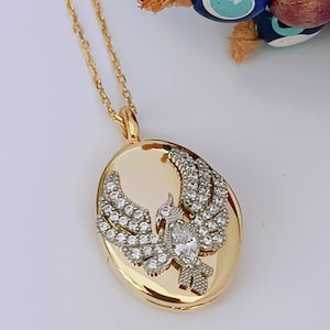 May include: Gold-toned necklace with an oval pendant featuring a detailed bird design embellished with clear crystals. The pendant hangs from a delicate chain. The background includes blue and brown beads.