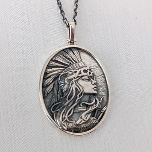 May include: A silver necklace with an oval pendant featuring a detailed relief of a woman wearing a headdress with feathers. The woman is looking to the right and has long hair.