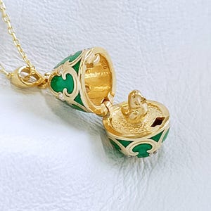 Könnte beinhalten: Eine goldfarbene Halskette mit einem aufklappbaren, eiförmigen Medaillon. Das Medaillon hat grüne Emaille-Details und eine kleine goldene Figur im Inneren. Das Medaillon ist geöffnet und gibt den Blick ins Innere frei. Die Kette ist filigran.