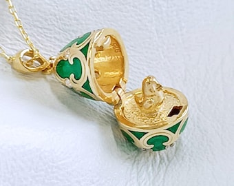 Pode incluir: Um colar com pingente dourado com um medalhão articulado em forma de ovo. O medalhão tem detalhes em esmalte verde e uma pequena figura dourada no interior. O medalhão está aberto, revelando o interior. A corrente é delicada.