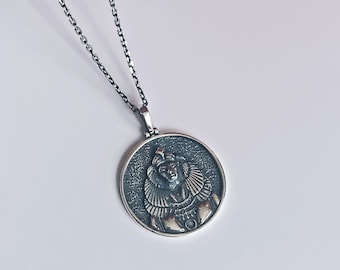 May include: A silver chain necklace with a round pendant featuring a detailed image of a woman's face. The pendant has a textured surface and a raised design.