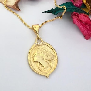 May include: A gold chain necklace with a pendant featuring a detailed profile of a woman's face. The pendant is oval-shaped and has a textured surface. The chain is delicate and has a simple design.