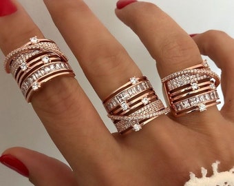 Puede incluir: Tres anillos de oro rosa con piedras blancas se muestran en una mano con esmalte de uñas rojo. Los anillos presentan un diseño en capas con piedras preciosas de diferentes tamaños y formas, incluyendo cortes baguette y redondos. Estos anillos son una pieza de joyería llamativa.