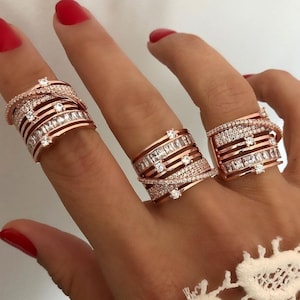 Könnte beinhalten: Drei roségoldene Ringe mit weißen Edelsteinen werden auf einer Hand mit rotem Nagellack gezeigt. Die Ringe haben ein geschichtetes Design mit verschiedenen Edelsteinformen, darunter Baguette- und Rundschliffe. Diese Ringe sind ein auffälliges Schmuckstück.