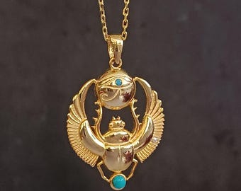 May include: Gold-colored pendant necklace featuring an Egyptian scarab beetle design. The pendant has a turquoise stone and an eye symbol. The necklace is on a gold chain.