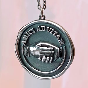 May include: A silver-colored pendant necklace featuring a circular charm with the Latin phrase "AMICI AD VITAM" and a depiction of two clasped hands. The pendant hangs from a delicate chain.