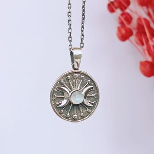 May include: A silver pendant necklace featuring a circular pendant with a triple moon design, a central stone, and radiating lines. The pendant hangs from a delicate chain. The background is a soft white with red floral accents.