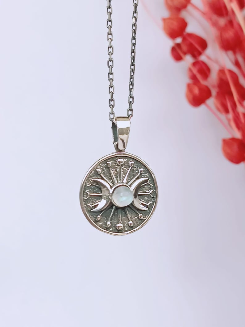 May include: A silver pendant necklace featuring a circular pendant with a triple moon design, a central stone, and radiating lines. The pendant hangs from a delicate chain. The background is a soft white with red floral accents.