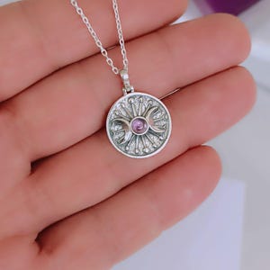 May include: A silver pendant necklace featuring a circular charm with a central purple gemstone. The charm has a detailed design with crescent moons and sunburst elements. The necklace is on a delicate chain.