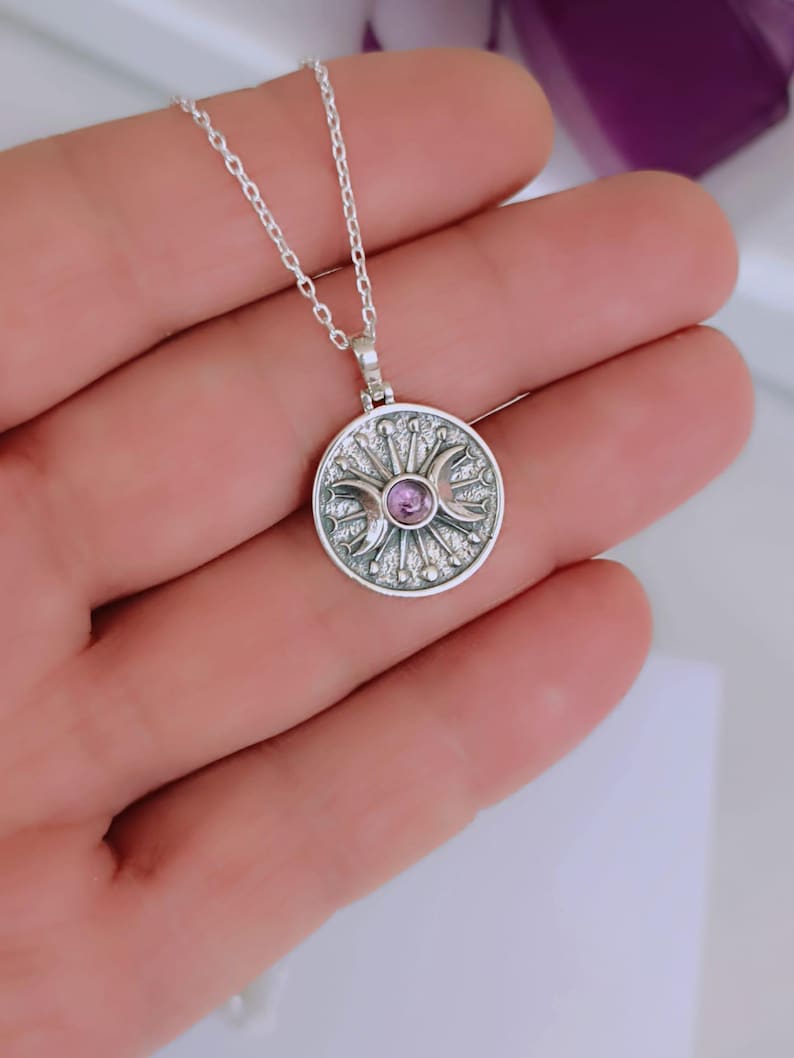 May include: A silver pendant necklace featuring a circular charm with a central purple gemstone. The charm has a detailed design with crescent moons and sunburst elements. The necklace is on a delicate chain.