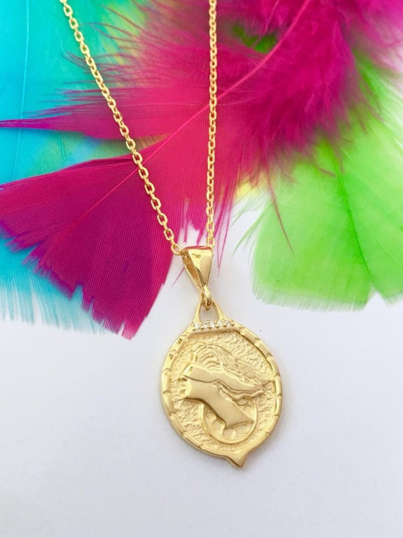 K&ouml;nnte beinhalten: Eine goldene Kettenkette mit einem runden Anh&auml;nger, der ein detailliertes Profil eines Frauenkopfes mit Heiligenschein zeigt. Der Anh&auml;nger ist mit kleinen, klaren Steinen am Rand verziert.