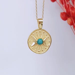 May include: A gold-colored pendant necklace featuring a circular pendant with a turquoise stone center. The pendant has a textured design with crescent moon shapes and sunburst details. The necklace chain is also gold-colored.