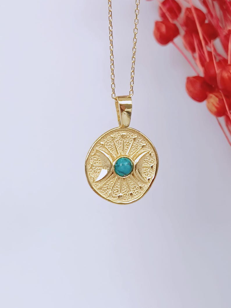 May include: A gold-colored pendant necklace featuring a circular pendant with a turquoise stone center. The pendant has a textured design with crescent moon shapes and sunburst details. The necklace chain is also gold-colored.
