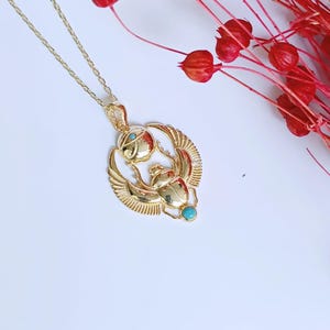 May include: Gold-colored pendant necklace featuring an Egyptian scarab beetle design with turquoise accents. The pendant has detailed wings and a circular element above the beetle. The necklace is on a white surface with red floral accents.