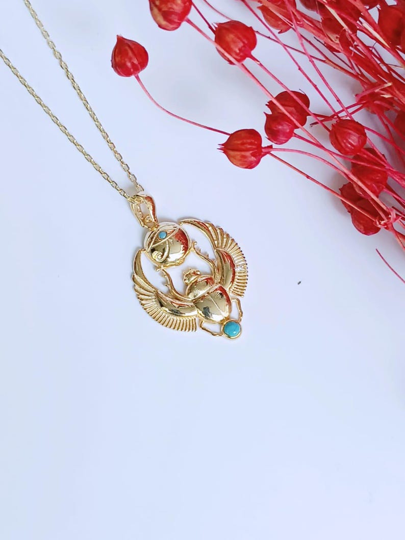 May include: Gold-colored pendant necklace featuring an Egyptian scarab beetle design with turquoise accents. The pendant has detailed wings and a circular element above the beetle. The necklace is on a white surface with red floral accents.