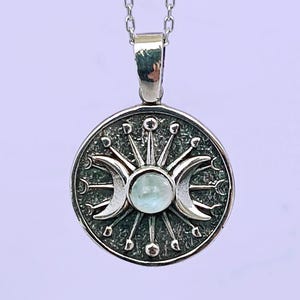 May include: A silver-toned pendant necklace featuring a circular pendant with a celestial design. The pendant showcases a central light blue gemstone, flanked by crescent moons and sunburst details. The necklace has a delicate chain.