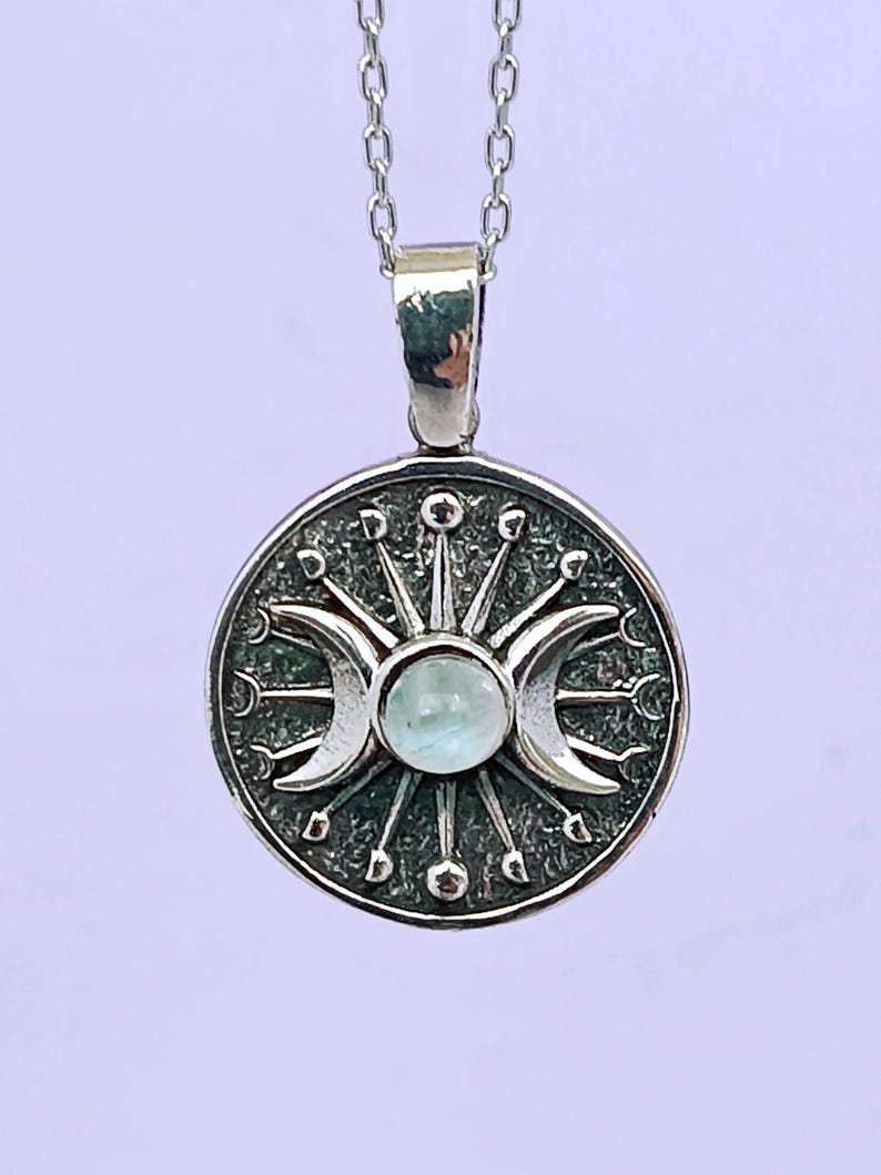 May include: A silver-toned pendant necklace featuring a circular pendant with a celestial design. The pendant showcases a central light blue gemstone, flanked by crescent moons and sunburst details. The necklace has a delicate chain.