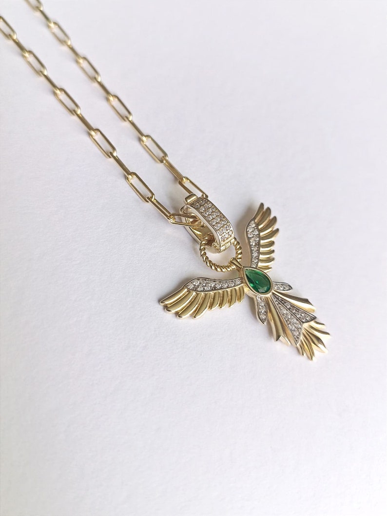 Pode incluir: Um colar de corrente de ouro com um pingente que apresenta um anjo estilizado com asas, uma gema verde e pedras brilhantes transparentes.