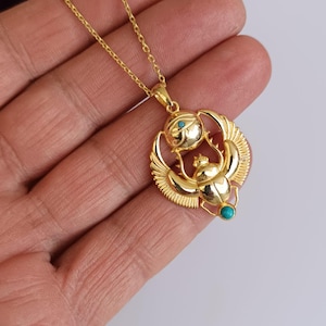 May include: Gold-colored pendant necklace featuring an Egyptian scarab beetle design with turquoise accents. The pendant has a detailed scarab beetle with outstretched wings, a sun disc, and a delicate chain.