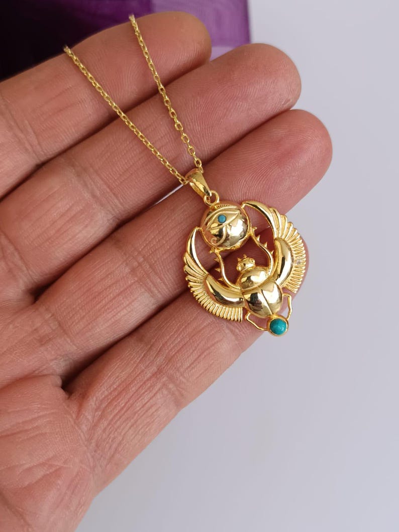 May include: Gold-colored pendant necklace featuring an Egyptian scarab beetle design with turquoise accents. The pendant has a detailed scarab beetle with outstretched wings, a sun disc, and a delicate chain.