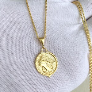 K&ouml;nnte beinhalten: Eine goldene Kettenkette mit einem runden Anh&auml;nger, der ein detailliertes Profil eines Frauenkopfes zeigt. Der Anh&auml;nger hat eine strukturierte Oberfl&auml;che und ist mit kleinen, klaren Steinen verziert.