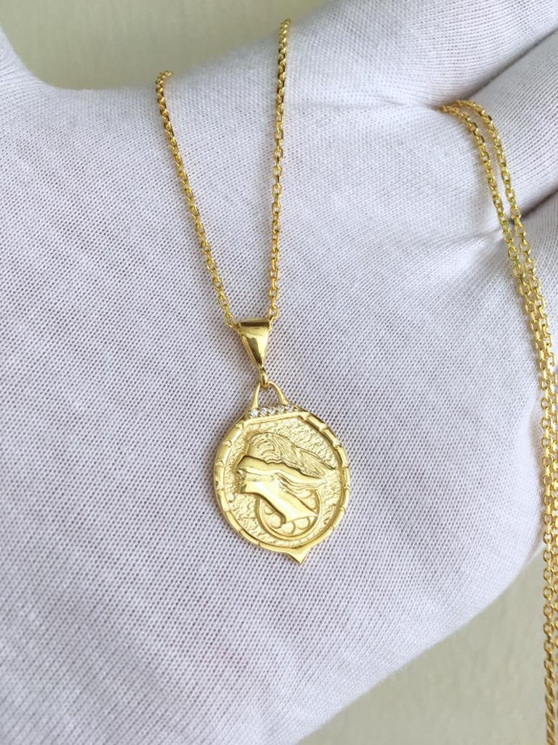 K&ouml;nnte beinhalten: Eine goldene Kettenkette mit einem runden Anh&auml;nger, der ein detailliertes Profil eines Frauenkopfes zeigt. Der Anh&auml;nger hat eine strukturierte Oberfl&auml;che und ist mit kleinen, klaren Steinen verziert.